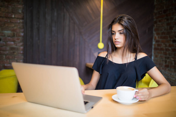 Jeune femme assise dans un café à une table en bois, buvant du café et utilisant un smartphone. Sur la table se trouve un ordinateur portable. Fille naviguant sur Internet, bavarder, blogging
. - Photo, image