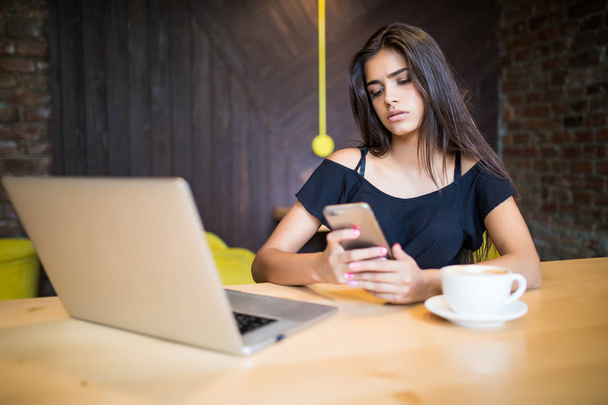 attraktive Freiberuflerinnen halten ihr Smartphone in der Hand, während sie am Holztisch vor dem offenen Computer in einem modernen Café sitzen. junge kreative Frau arbeitet beim Frühstück am Laptop - Foto, Bild