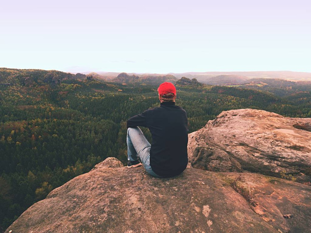 Tramp muž na skále. Turista v červené čepici, černý svetr a džíny sedí na ostré horské skále nad údolím - Fotografie, Obrázek