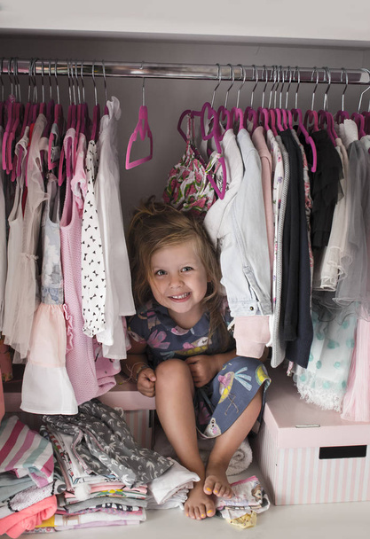 Красивая маленькая девочка сидит в шкафу и улыбается. Девичья одежда на вешалках в шкафу
. - Фото, изображение