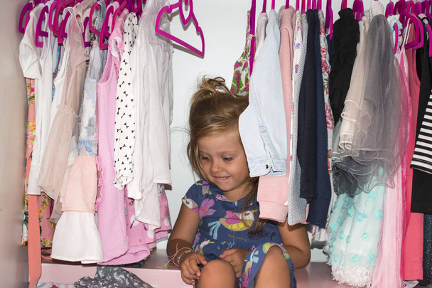 Hermosa niña sentada en su armario y sonriendo. Ropa de niña en perchas en el armario
. - Foto, Imagen