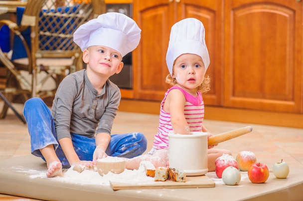 Dva sourozenci - chlapec a dívka - šéfkuchaře klobouky sedí na kuchyni. - Fotografie, Obrázek