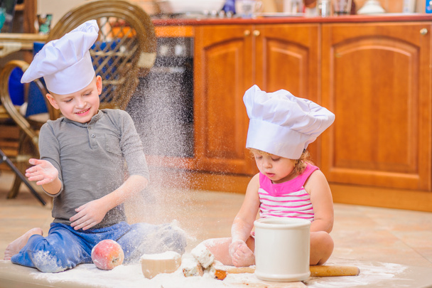 Dva sourozenci - chlapec a dívka - šéfkuchaře klobouky sedí na kuchyni. - Fotografie, Obrázek