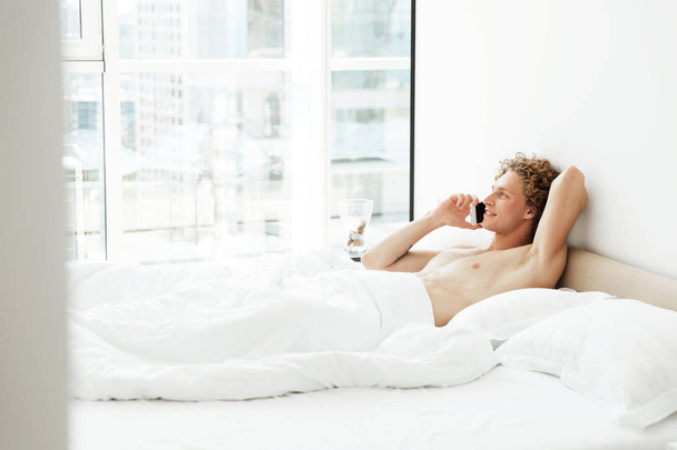 Cheerful man lies in bed at home talking by phone - Photo, image