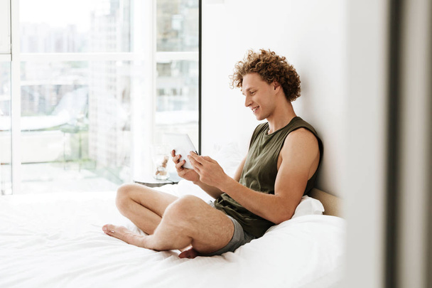 Happy man sitting on bed at home using tablet computer - Foto, Bild