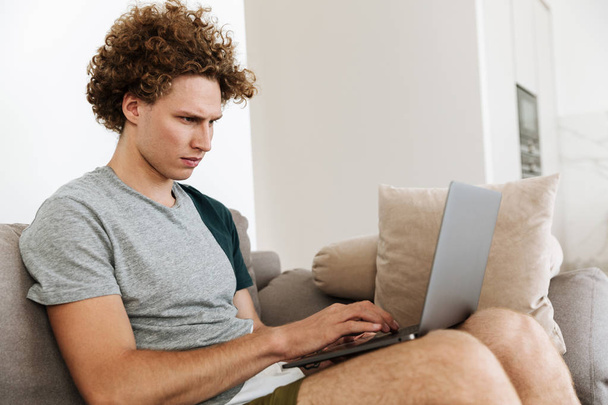 Красивий концентрований чоловік сидить на дивані, використовуючи ноутбук
 - Фото, зображення
