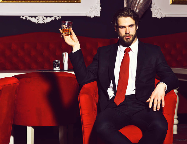 bearded man, businessman holding glass of whiskey in red chair - Foto, afbeelding