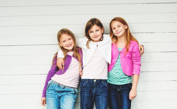 Group of 3 little girls standing outdoors against white wooden background, wearing denim jeans and colorful jackets - Fotó, kép