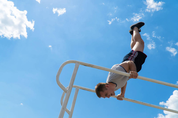 The guy is standing on his hands on the uneven bars. Workout outdoors. Blue sky on blackground - Фото, зображення