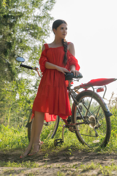 Kırmızı elbiseli bir genç kız bir bisiklet kırmızı şemsiye ile yakın duruyor. Arka planda bir orman var - Fotoğraf, Görsel