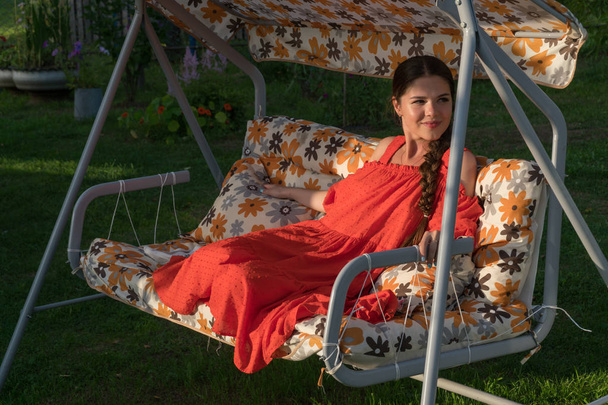A young girl in a red dress is resting on a country swing - Valokuva, kuva