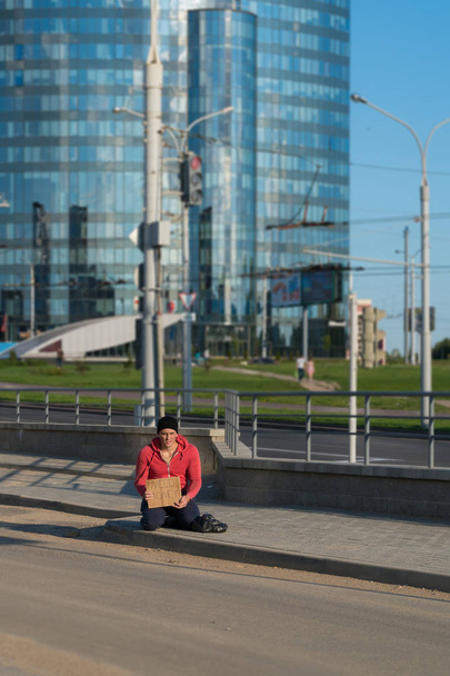 Бездомный сидит на тротуаре с картонкой и надписью: нужны деньги. На заднем плане бизнес-центр
 - Фото, изображение
