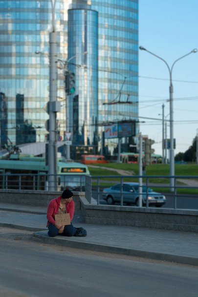 Bezdomnych facet siedzi na chodniku z kartonu i napis: potrzebują pieniędzy. W tle znajduje się centrum biznesowe - Zdjęcie, obraz