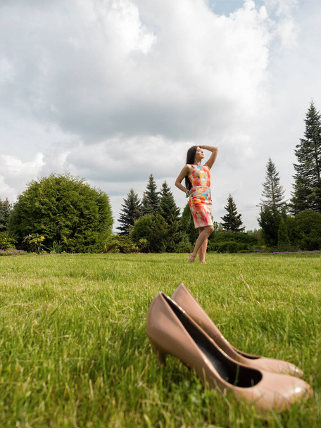 Elbiseli kıza ayakkabısını fırlatıp aldı ve kendini zevk oldu. Kız çıplak ayakla çimlerde yürüyor. Ön planda bej Ayakkabı - Fotoğraf, Görsel