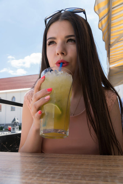 A girl is drinking a tropical mojito in a summer cafe. A girl with glasses, a pink dress and bright red lips - Foto, Imagem
