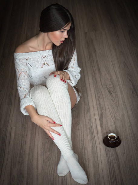 Tyttö, jolla on valkoinen pusero ja sukat, istuu jalopuulattialla kahvin kanssa.
 - Valokuva, kuva