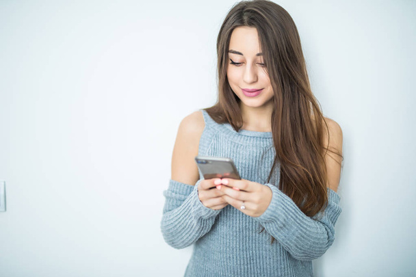 Enviar mensajes a un amigo. Atractiva joven mujer sosteniendo el teléfono inteligente y mirándolo con sonrisa mientras está de pie sobre un fondo gris
 - Foto, imagen