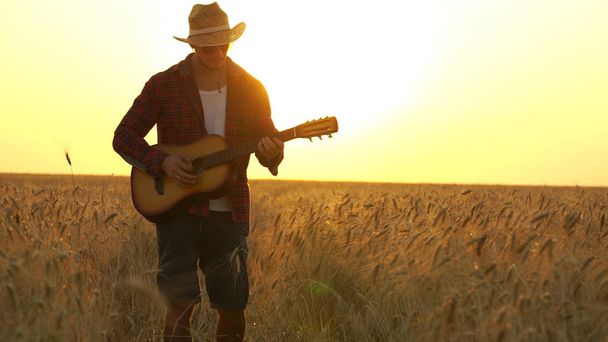joven camina por el campo de trigo dorado en los rayos del atardecer y toca la guitarra
. - Foto, Imagen