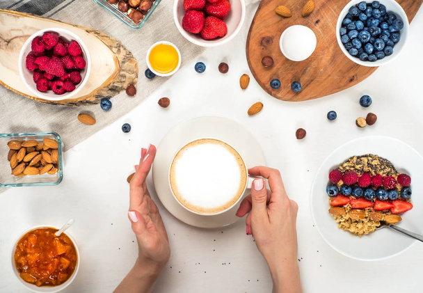 Good morning - healthy breakfast background with oatmeal coffee, berries, egg, nuts. Coffee, hands, hold, cup. White wooden food background, top view - Foto, Bild