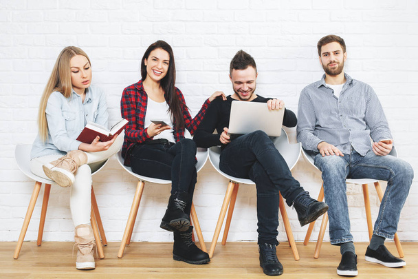 Young businessmen and women sitting on chairs, reading book and using laptop in studio with white brick wall and wooden floor. Partnership concept  - Foto, afbeelding