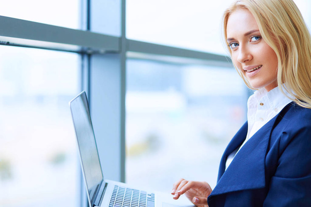 Businesswoman standing against office window holding laptop - 写真・画像