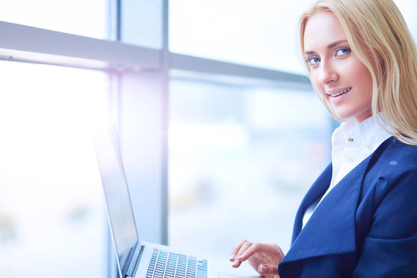 Businesswoman standing against office window holding laptop - Foto, afbeelding