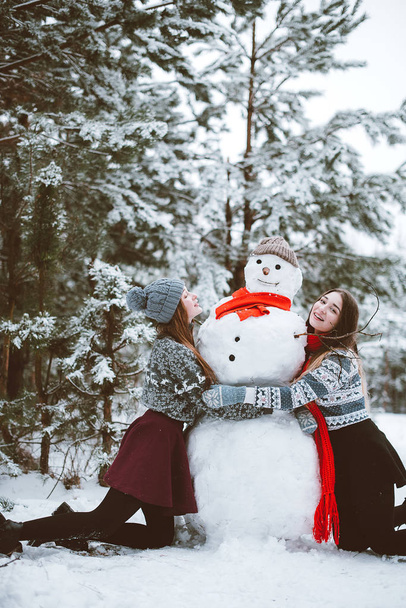 Two young teenage hipster girl friends together.Close up fashion portrait of two sisters hugs and having fun, make snowman in winter time forest, wearing sweaters and scarfs,best friends couple outdoo - Φωτογραφία, εικόνα