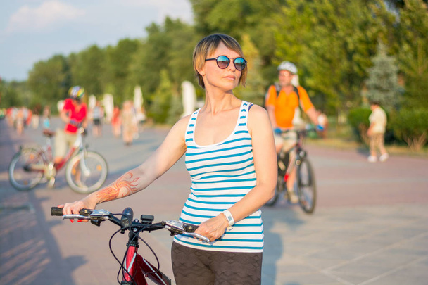 Μια νεαρή γυναίκα σε γυαλιά ηλίου στέκεται με ένα ποδήλατο για το promena - Φωτογραφία, εικόνα