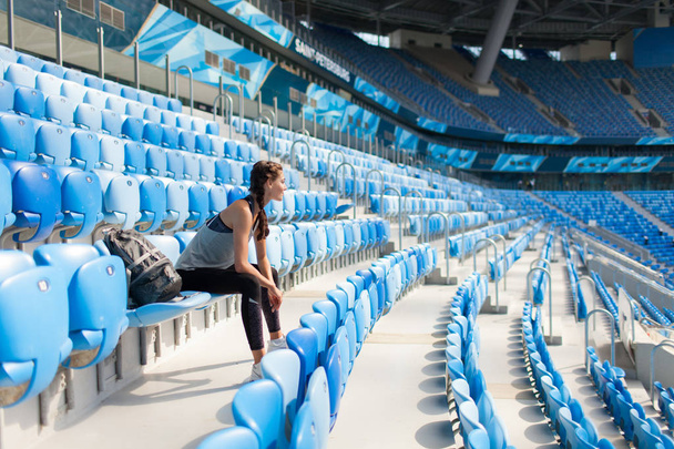 Sexy girl posing at stadium with the backpack. Fitness girl with a sports figure in black leggings sitting on the seat in the stadium. Sport woman posing at stadium. Girl smiling after sport day - Фото, изображение