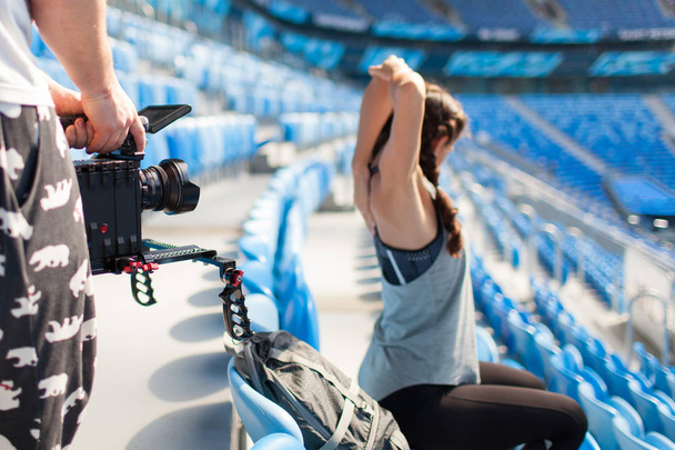 El videógrafo dispara en una cámara profesional como una chica deportiva calentándose, sentada en el estadio
 - Foto, Imagen
