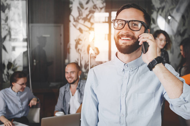 Photo close-up young smiling bearded businessman in glasses speaking on smartphone on background coworking meeting business team. Man talking on phone with creative director. Intentional lens flares - Foto, Imagem