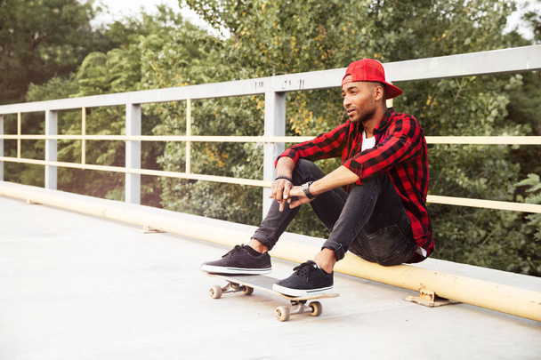 Young dark skinned man sitting with his skateboard - Фото, изображение