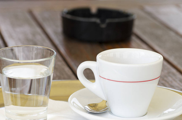Close-up foto van een kopje koffie op een schotel met een lepel naast een glas water en een asbak met een onscherpe achtergrond  - Foto, afbeelding
