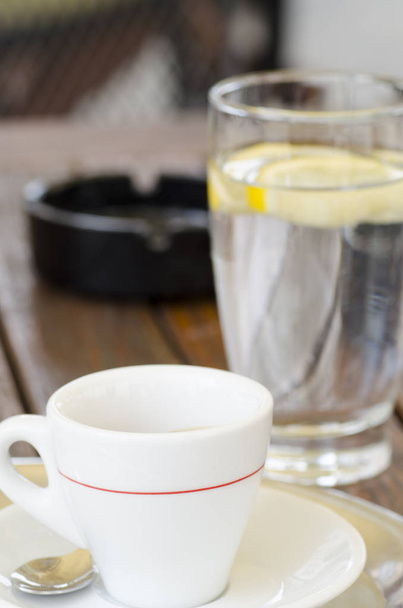 Detail fotografie šálek kávy na talířek se lžičkou vedle sklenice vody a popelník s rozmazané pozadí - Fotografie, Obrázek