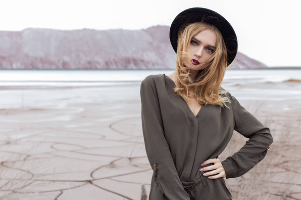 Beautiful, sexy blond girl posing in the mountains on a cracked ground in a black hat with bright make-up - Φωτογραφία, εικόνα