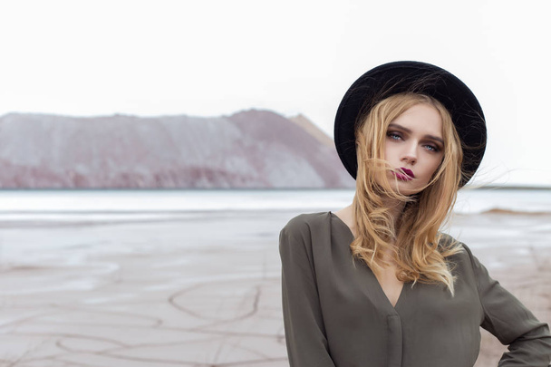 Beautiful, sexy blond girl posing in the mountains on a cracked ground in a black hat with bright make-up - Photo, Image