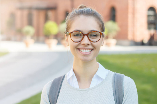 Closeup portrait of good-looking happy college student girl, wearing big fashionable glasses and grey backpack, her hair tied in bun, smiling friendly at camera, feeling satisfied and joyful. - Foto, Imagen