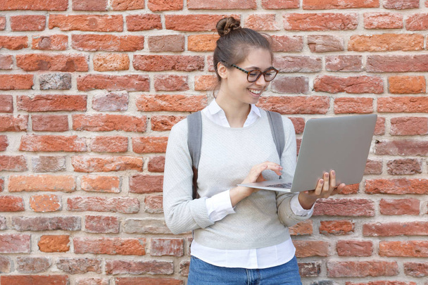 Closeup photo of young European female standing against red brick background smiling happily while holding laptop in hands, using touchpad to browse in web and reply to messages from friends. - Фото, изображение