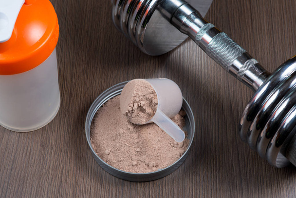 Whey protein in a measuring bucket with a metal dumbbell and shaker on a wooden background - Foto, afbeelding
