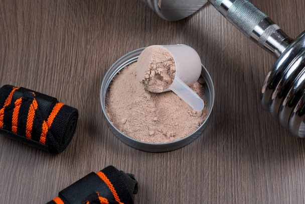 Whey protein in a measuring bucket with a metal dumbbell on a wooden background - Φωτογραφία, εικόνα