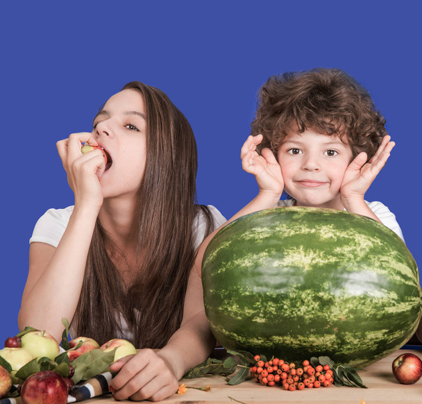 Brother and sister sitting at a table. Wait a delicious watermelon. Sister eating apple. Brother smiling and looking at the camera. Blue background. Close-up. - Zdjęcie, obraz