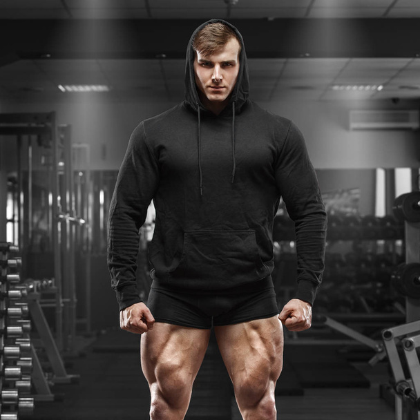 Muscular man with muscle legs in gym. Strong male in black hoodie with big quads - Photo, Image