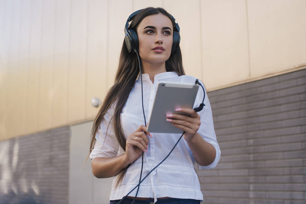 A very beautiful girl dressed in black big headphones smiles happily. brunette girl runs and listens to music. Sound quality in the headphones. The girl holds the iPad tablet in her hand - Photo, Image