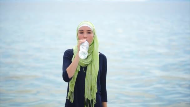 Μια νεαρή γυναίκα σε ένα hijab πίνει μεταλλικό νερό από το μπουκάλι σε μια καυτή ημέρα σε εξωτερικούς χώρους - Πλάνα, βίντεο