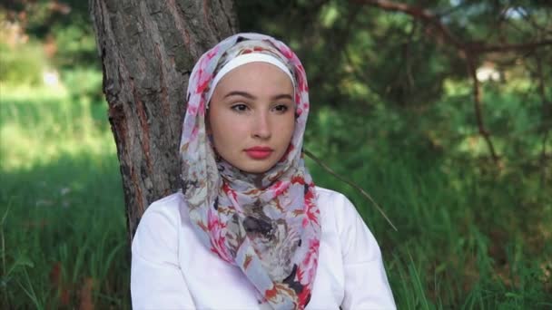 Žena v muslimský šátek sedí na trávě vedle stromu v parku - Záběry, video