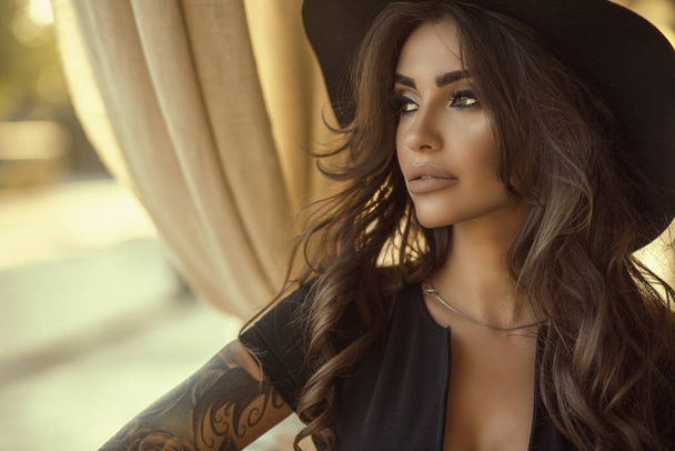 Close up portrait of beautiful tanned glam tattooed model with long wavy hair wearing black dress and wide brimmed hat looking aside - Foto, Imagen