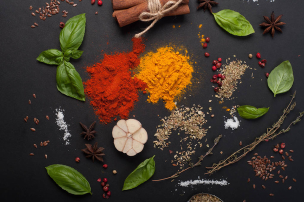 Bright spices, seasonings and herbs (turmeric, paprika, pink pepper, cinnamon, basil, thyme, anise and garlic) on a dark board - Foto, imagen
