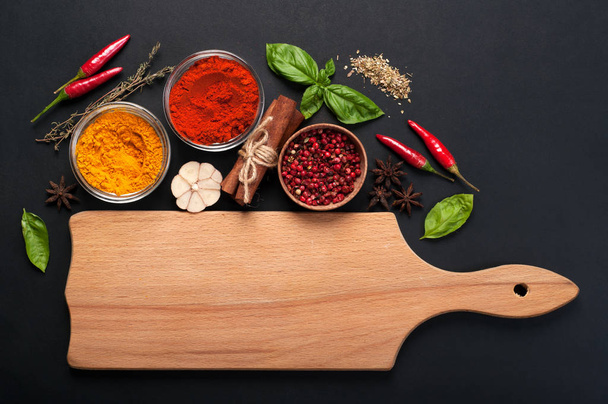 Bright spices, seasonings and herbs (turmeric, paprika, pink pepper, cinnamon, basil, thyme, anise and garlic) on a dark background. Near the light brown wooden cutting board (space for text) - Foto, Imagen