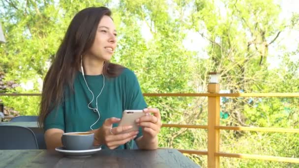 Nainen, joka käyttää sovellusta älypuhelimella kahvilassa juomassa kahvia hymyillen ja kutomalla matkapuhelimella. Kaunis nuori nainen ammattilainen matkapuhelimella
. - Materiaali, video