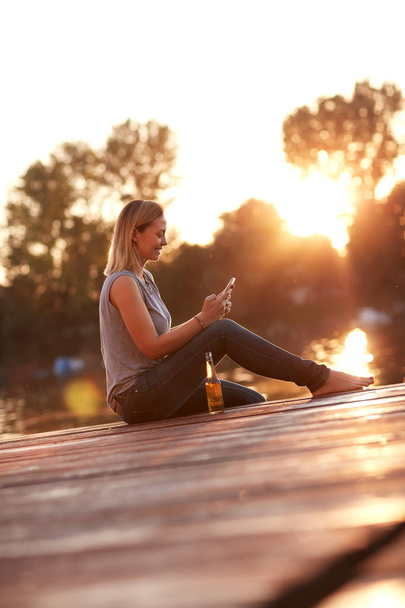 Girl sitting on dock at sunset and looking at phone  - Foto, Bild